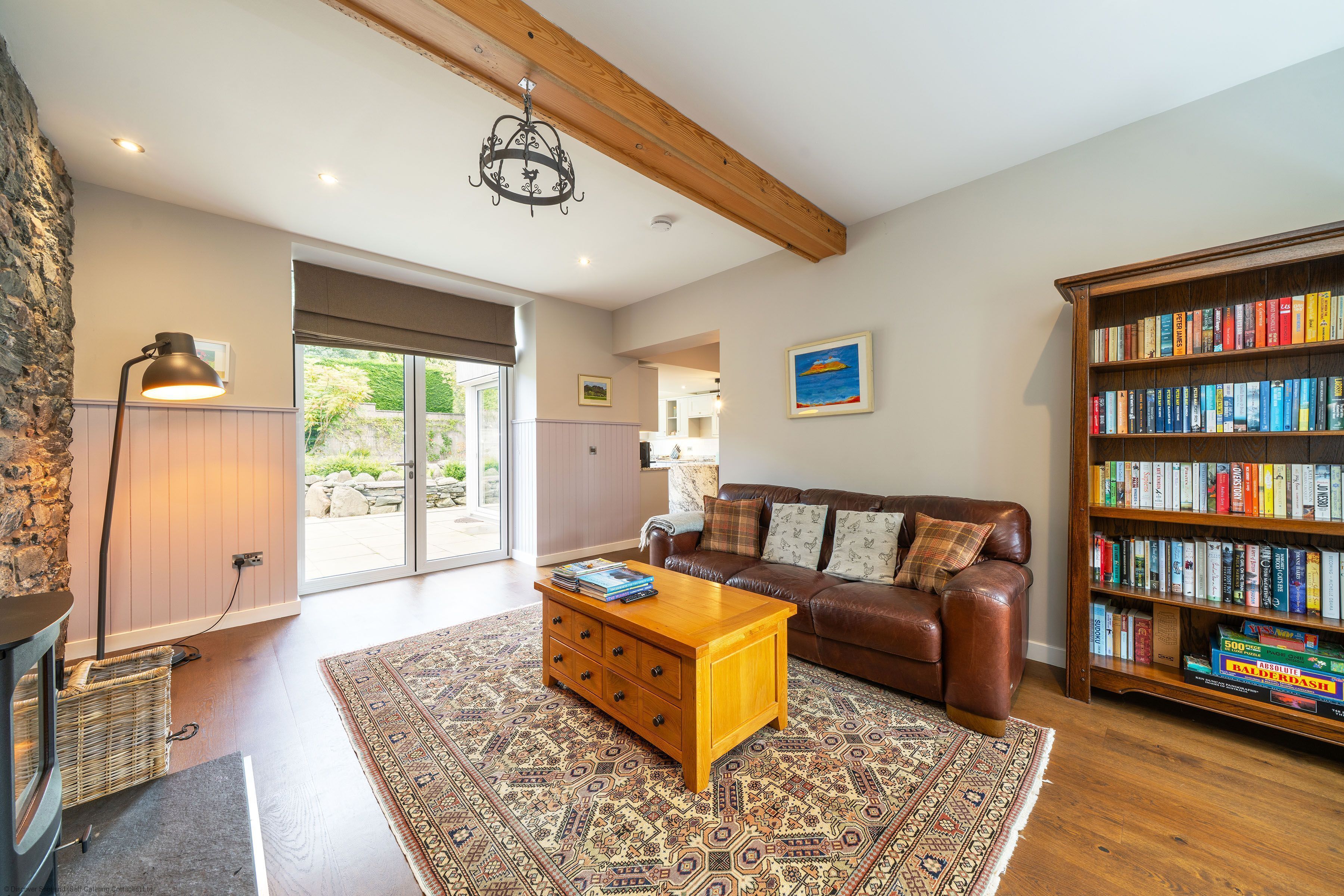 A spacious living room with a leather sofa, a wooden coffee table, a patterned rug, a bookshelf, and a large window.
