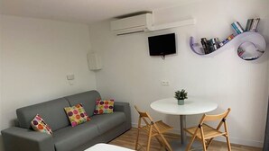 A modern room with a grey sofa, a round table with two chairs, a flat-screen TV, and a bookshelf with books and decorative items. - Hôtel Le Thimothée (Saint-Raphaël)