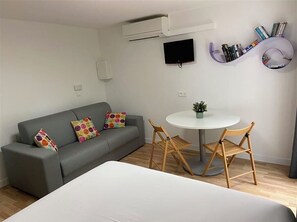 Een moderne kamer met een grijze bank, een ronde tafel met twee stoelen, een flatscreen televisie en een boekenkast met boeken en decoratieve objecten.