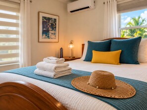 Quarto de hotel com cama, pilha de toalhas, chapéu e pintura na parede.