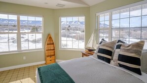 A bedroom with a large bed, two windows with a snowy landscape view, a wooden shelf, and a sailboat model on a nightstand.