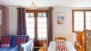 Een kamer met een bank, een eettafel met tafelkleed, een houten stoel, een houten deur en een raam met gordijnen.