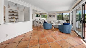 A spacious living area with a dining table, two blue sofas, and a large window offering a view of greenery. - The Polo Lounge By The Sea 3BD w Large Patio (Carpinteria)