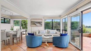 A spacious living room with a large sofa, two blue armchairs, a coffee table, and a dining area with a table and chairs. - The Polo Lounge By The Sea 3BD w Large Patio (Carpinteria)