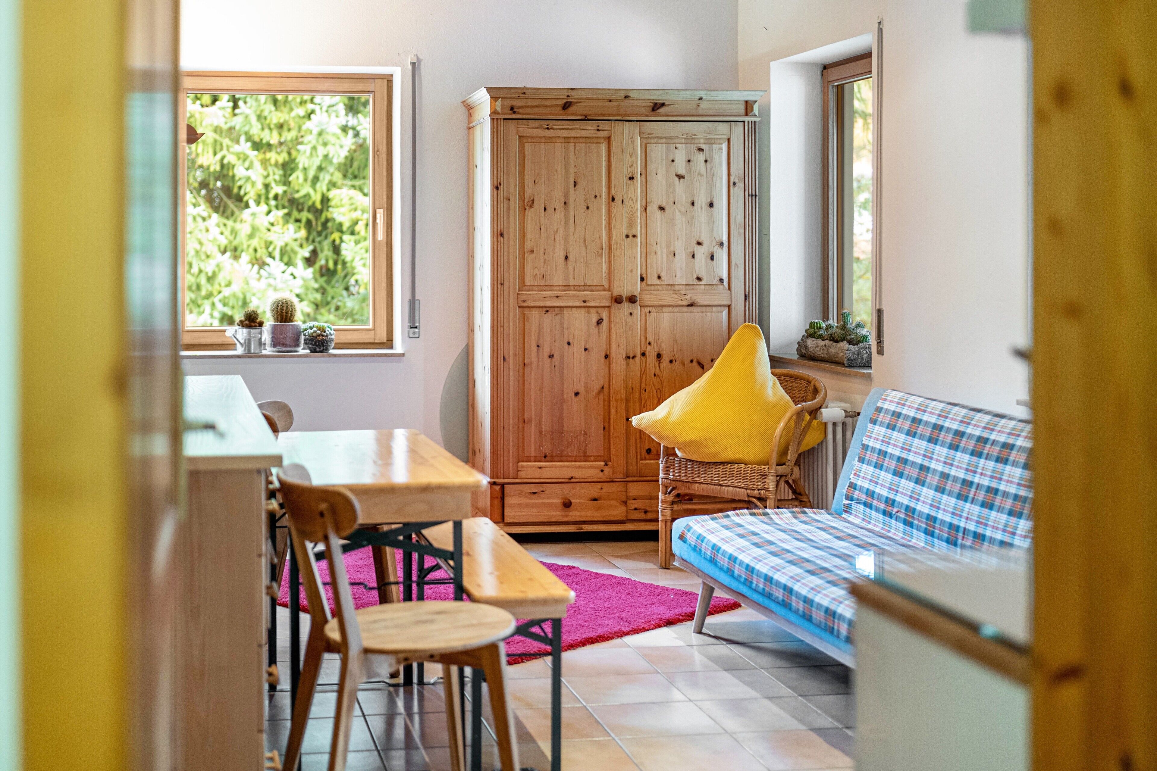 Una habitación con un armario de madera, una mesa de comedor con sillas, un sofá con funda escocesa y una ventana con plantas.