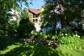 Une maison dotée d’un balcon, entourée d’une végétation luxuriante et d’un mur de pierre.
