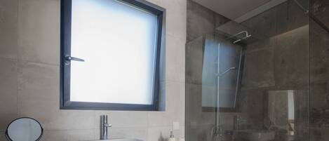 A modern bathroom with a large window, a round mirror, a white sink, and a glass shower enclosure.
