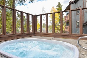 Ein Whirlpool mit einer Holzterrasse und Glasverkleidung, umgeben von Bäumen und einem Gebäude.