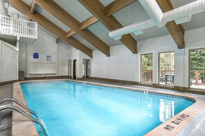 An indoor swimming pool with a clear view of the ceiling structure, a poolside area with a table and chairs, and a door leading to another room.