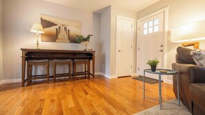 A living room with hardwood flooring, a brown leather couch, a coffee table, and a side table with a plant. There is a door with a window, a lamp, and a painting on the wall. - 8 mins to Lake Monroe • Grill • Firepit • By IU (bloomington)