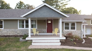 A two-story house with a stone foundation, a dark gray roof, and a red front door. There are two yellow Adirondack chairs on the front porch. - 8 mins to Lake Monroe • Grill • Firepit • By IU (bloomington)