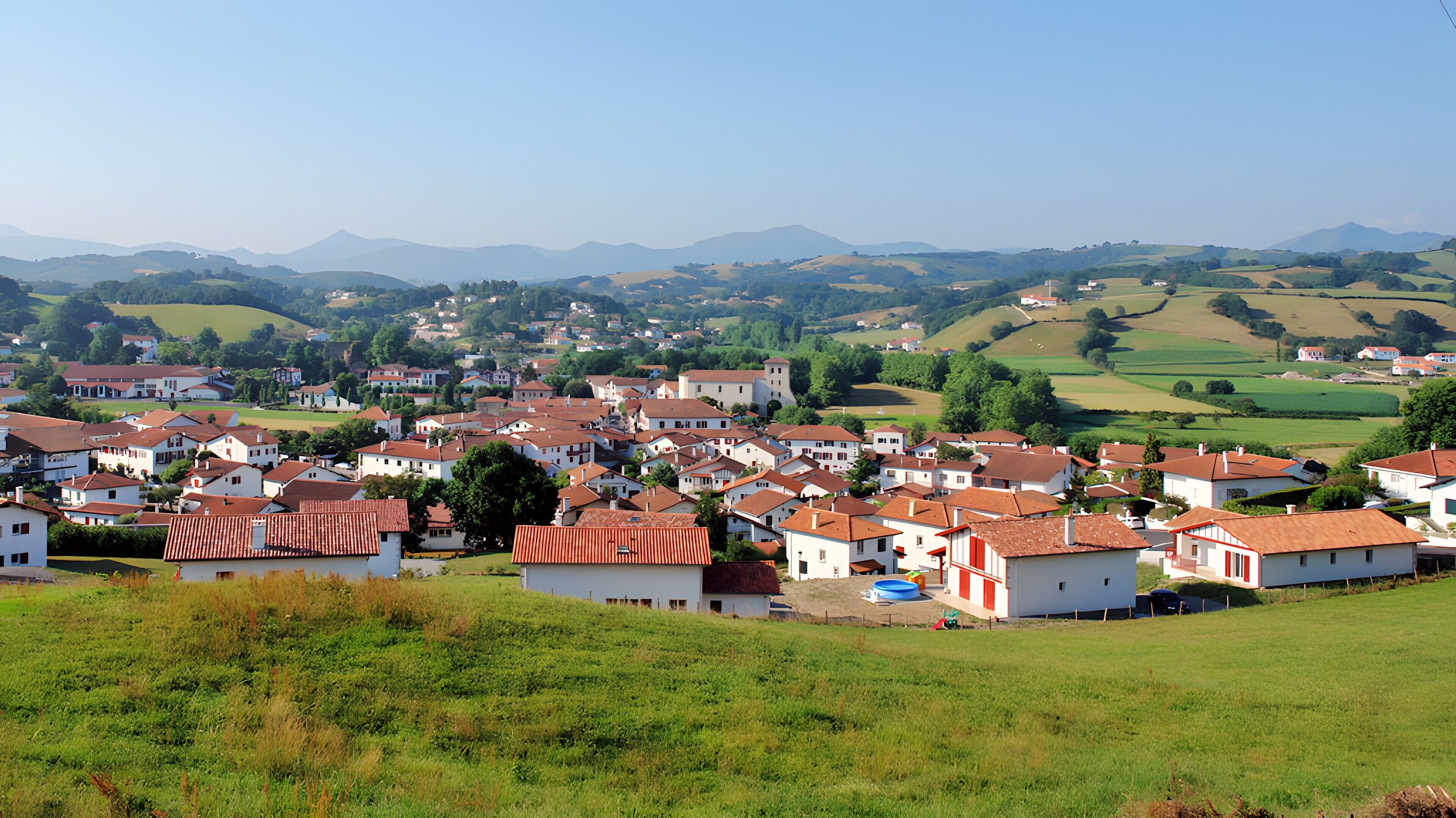 A village with white buildings and red roofs, surrounded by green fields and mountains.