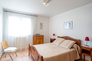 Een slaapkamer met een bed, een stoel, een houten nachtkastje, een rode lamp en een raam met gordijnen.