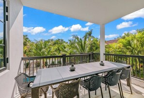 A covered outdoor dining area with a table and chairs, overlooking a tropical landscape with palm trees. - New 8/8 bathroom beach house! Heated pool and spa! (Sarasota)