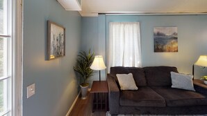 A living room with a brown sofa, two cushions, a side table with a lamp, a potted plant, and a painting on the wall. - 4297 Earney Rd, Unit 3, Woodstock (Woodstock)