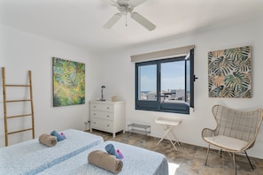 A bedroom with two beds, a window with a view, a ceiling fan, and artwork on the walls.