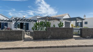 A modern building with a stone wall entrance, a covered patio area, and a clear blue sky. - Casa Bidasoa 35: Sea Views, Shared Pool & Wi-Fi in Playa Blanca (Playa Blanca)
