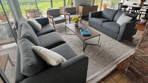 A spacious living room with a large sofa set, a coffee table, and a flat-screen TV. The room has large windows offering a view of a lush green landscape.