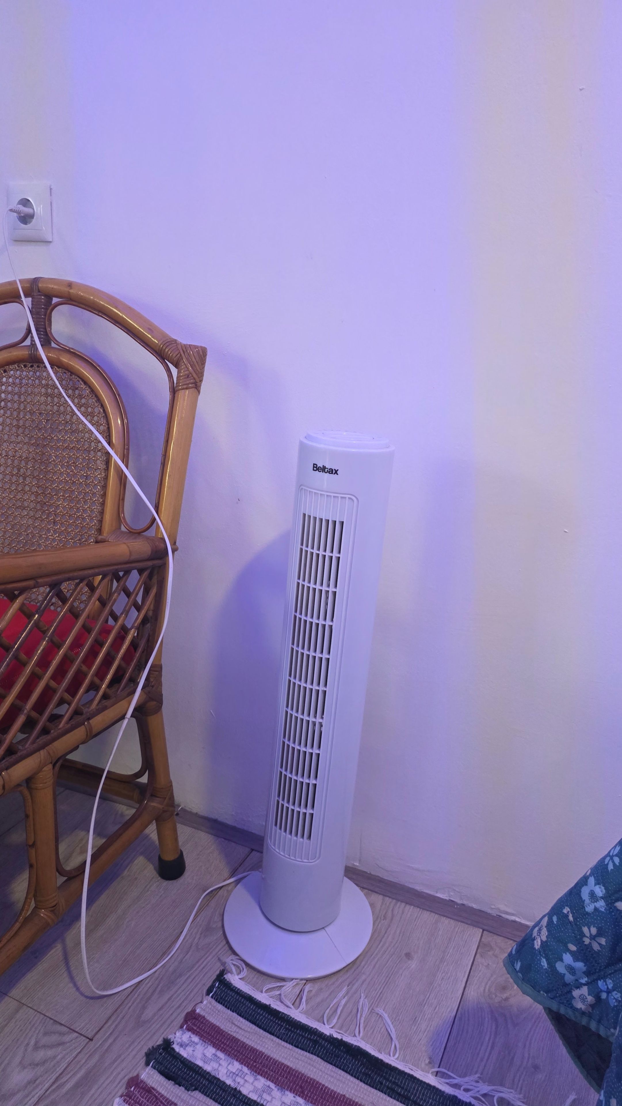 A white portable air conditioner with a cord plugged in, placed on a wooden floor next to a wicker chair and a patterned rug.