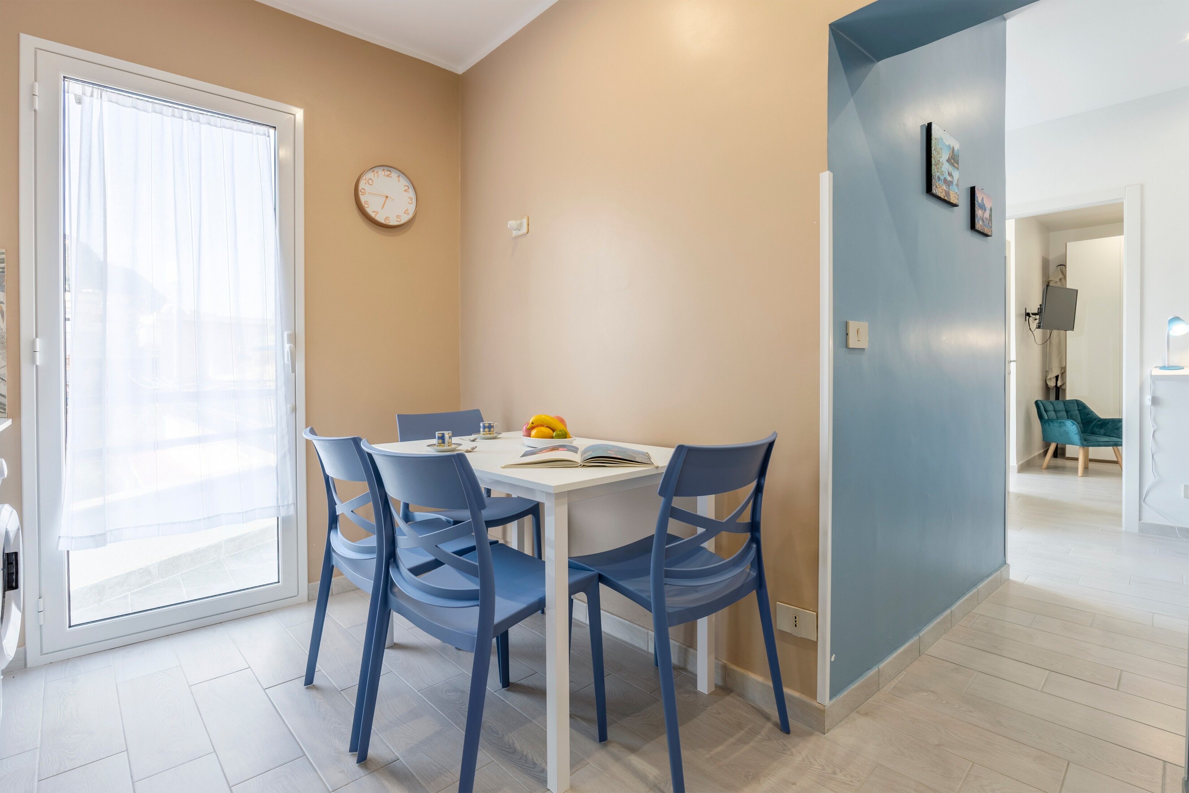 Un comedor con una mesa blanca, sillas azules, un reloj en la pared y una puerta que da a un balcón.