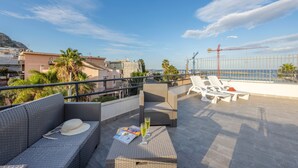 A rooftop terrace with wicker furniture, a table with a hat and glass, and a view of the sea and construction cranes. - La terrazza di porticello (Porticello)