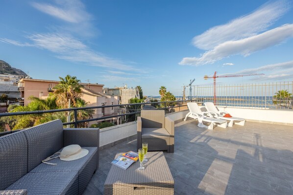 Une terrasse sur le toit, meublée de mobilier en osier, avec une table sur laquelle se trouvent un chapeau et un verre, et d’où l’on peut admirer la mer et les grues de chantier.