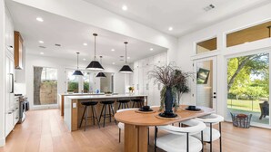 A modern kitchen with a central island, bar stools, pendant lighting, and a large window providing a view of the outdoors. - 7BR Estate - Pool on Golf Course - 4mi to Downtown (Austin)