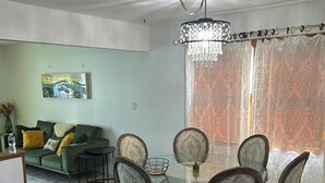A dining area with a glass table surrounded by patterned chairs, a chandelier, and a view of a living room with a sofa and a painting. - 2-bedroom townhome in charming South Milwaukee with AC (South Milwaukee)