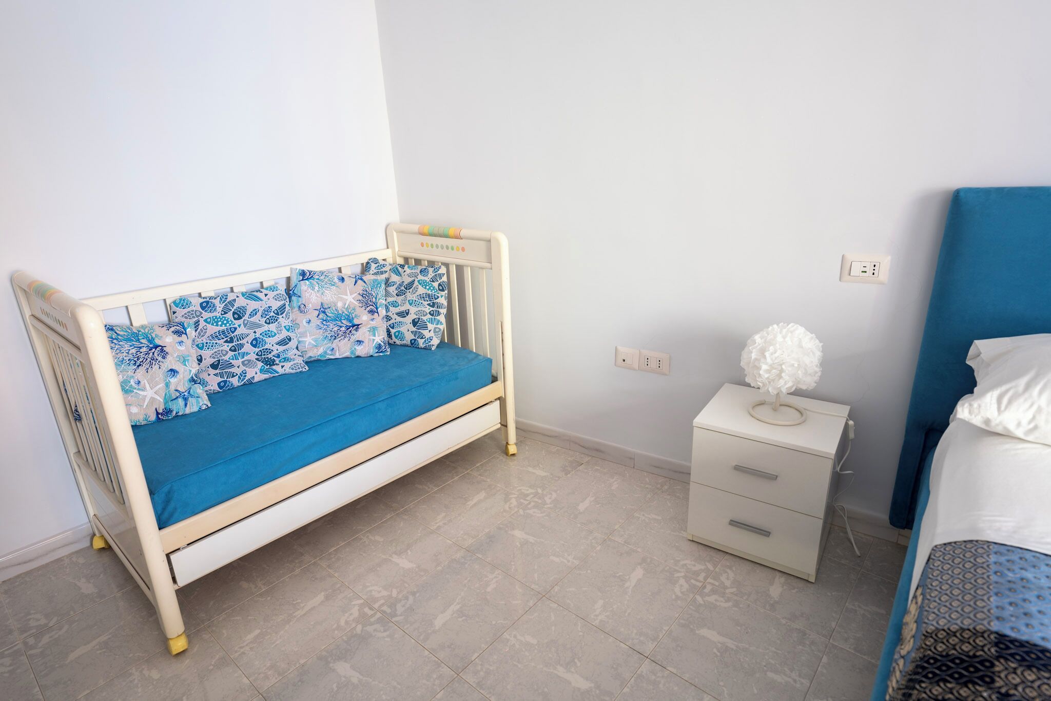 A child’s bed with a blue mattress and patterned pillows, a white nightstand with drawers, and a white wall socket.