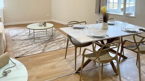 A modern dining area with a white table set for four, a round glass table with a plant, and a TV mounted on the wall displaying a scenic image. - Casa Luna – 1 Bedroom, 4 Guests, City Center, Fully Equipped (Metz)