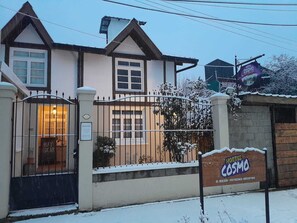 A two-story building with a gated entrance, a sign for Hostel Cosmos, and snow on the ground. - Hostel Cosmo (El Bolson)