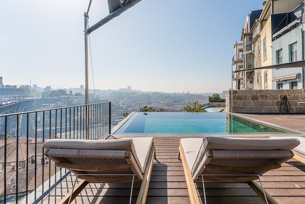 A rooftop infinity pool with two lounge chairs, a stone wall, and a view of the cityscape.