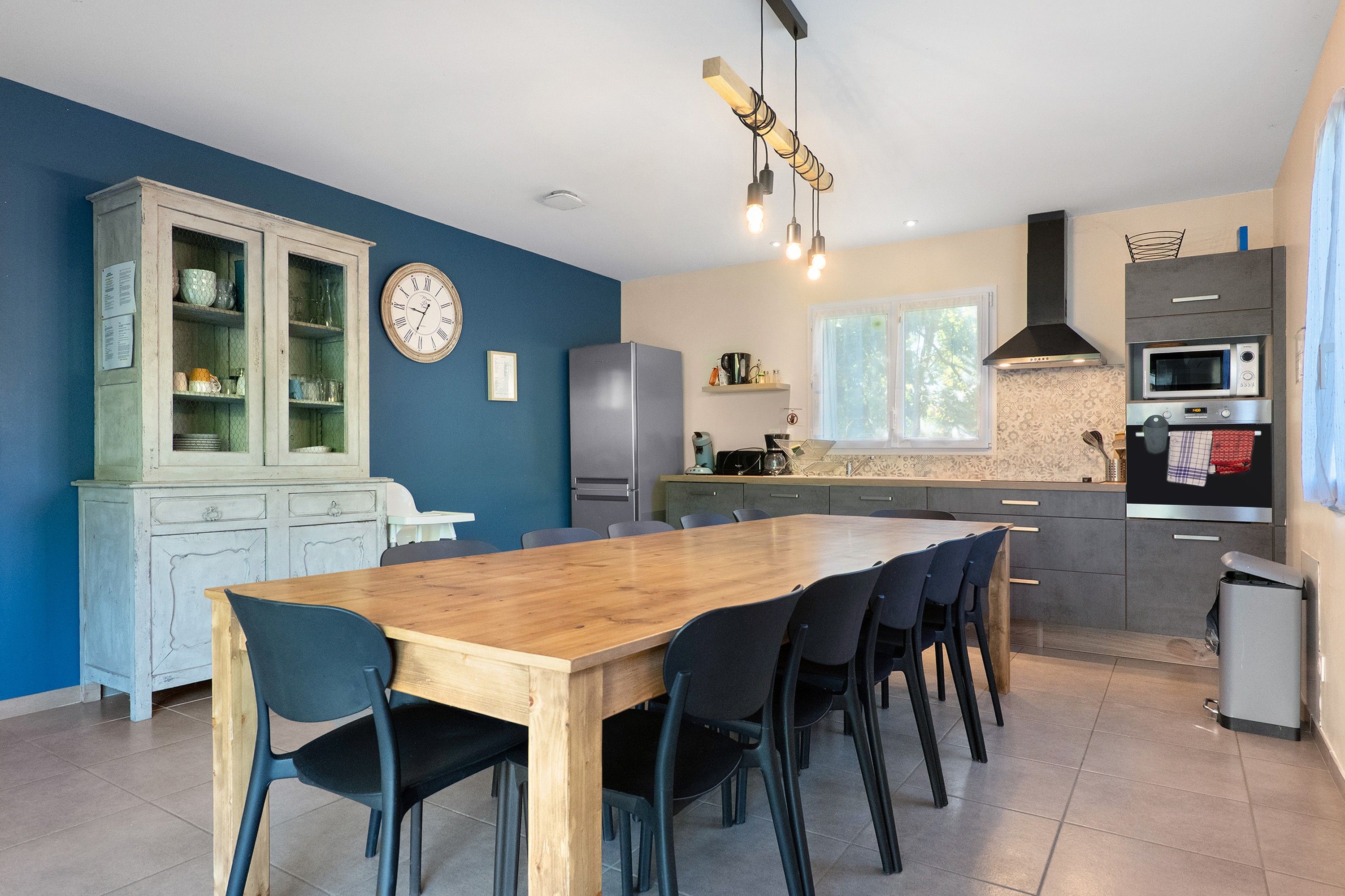 A modern kitchen with a dining area, featuring a wooden table and chairs, a built-in oven, and a clock on the wall.