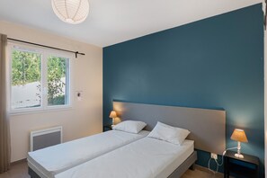 Quarto com cama, duas mesas de cabeceira com luminárias, janela com cortinas e radiador.