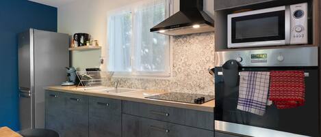 A modern kitchen with a stainless steel refrigerator, a microwave, and a built-in oven. There is a window with sheer curtains, a sink, and a countertop with various kitchen items.