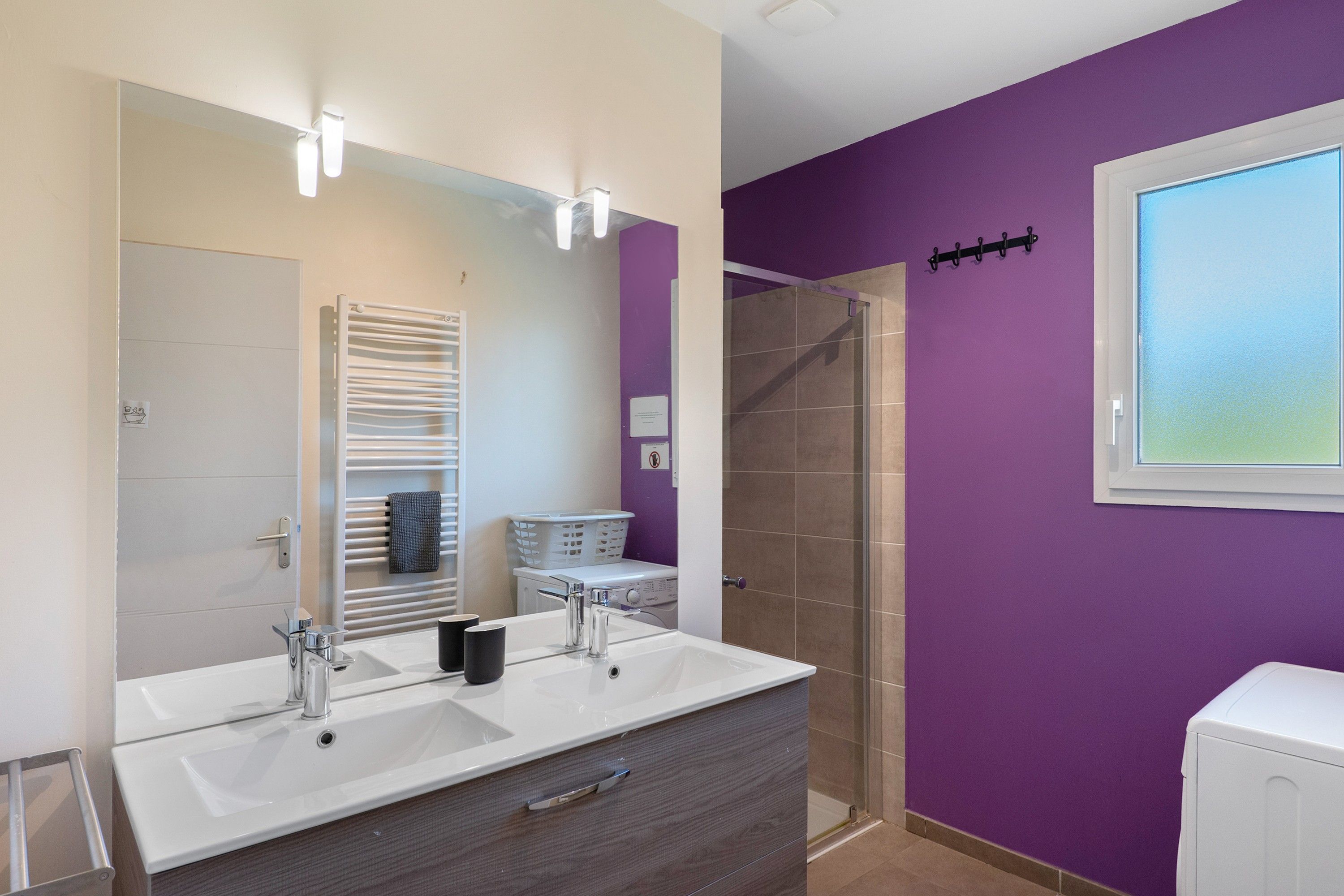 Une salle de bain avec un double lavabo, un miroir, un porte-serviettes, une douche et une fenêtre.