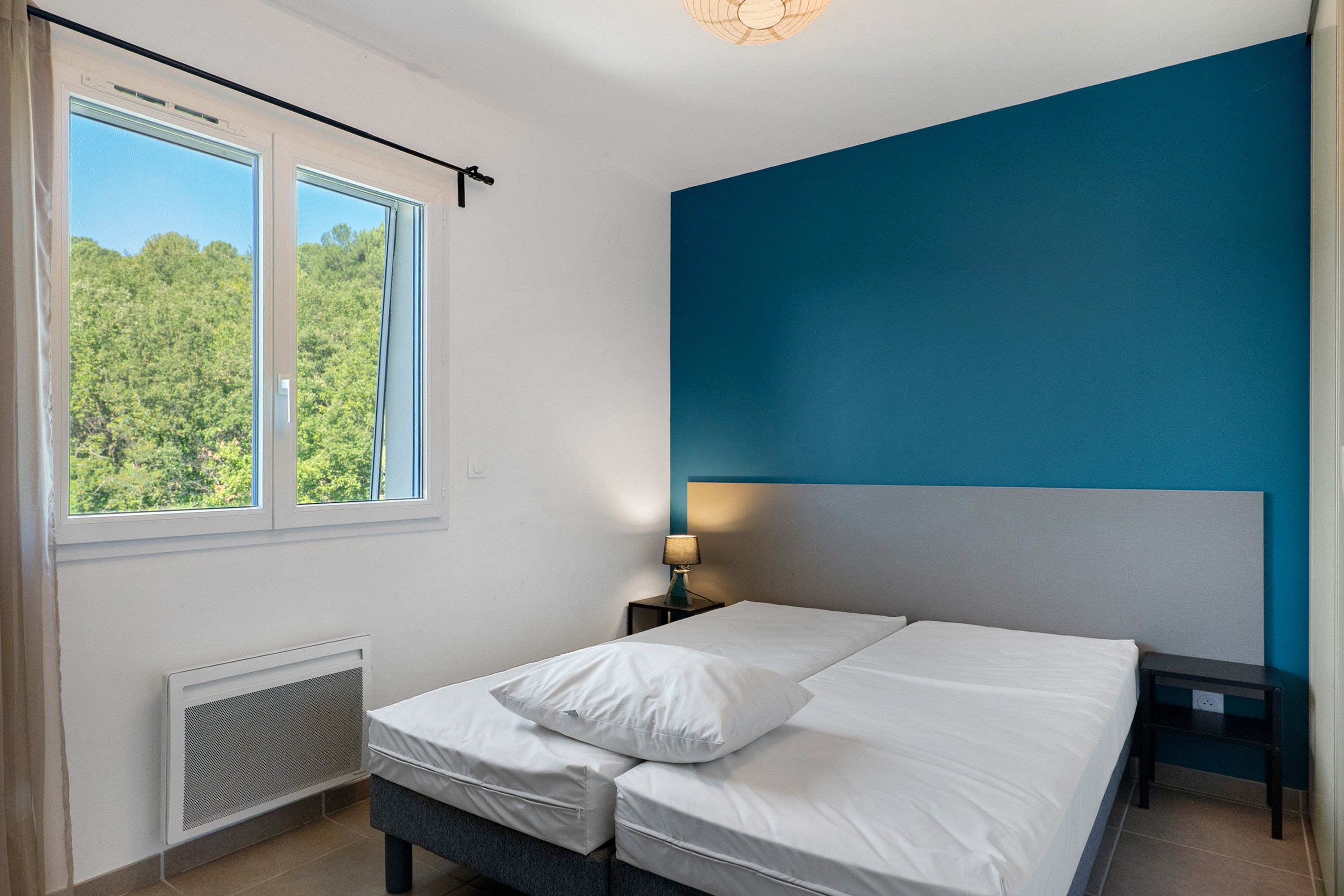 Een slaapkamer met een bed, een nachtkastje, een lamp en een raam met uitzicht op bomen.