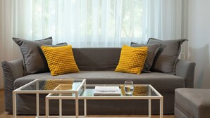 Un salon moderne avec un canapé gris, des coussins jaunes et gris, et une table basse en verre sur laquelle se trouvent un livre et un verre d’eau.