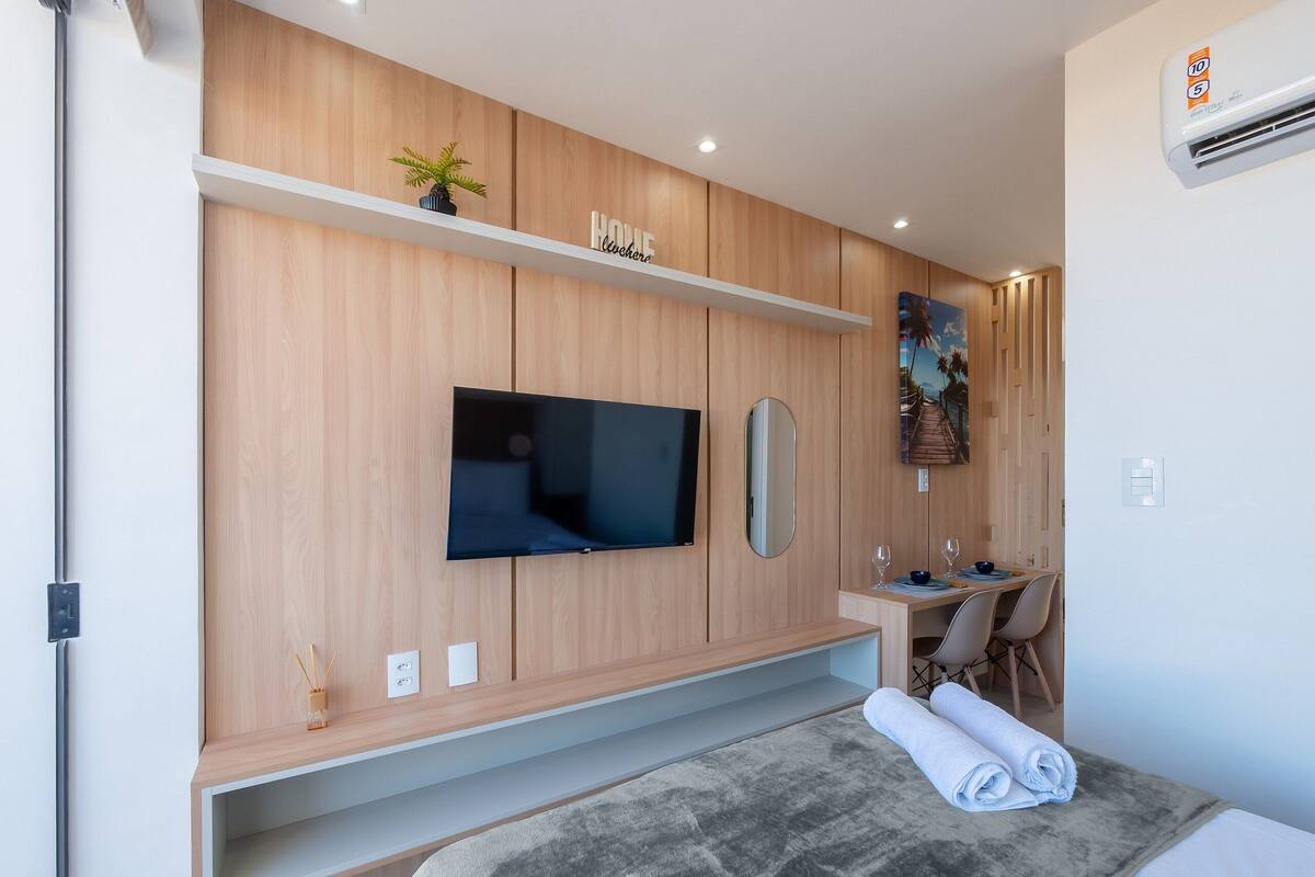 A modern hotel room with a flat-screen TV mounted on a wooden wall, a small plant on a shelf, a painting, and a neatly made bed with a rolled towel.