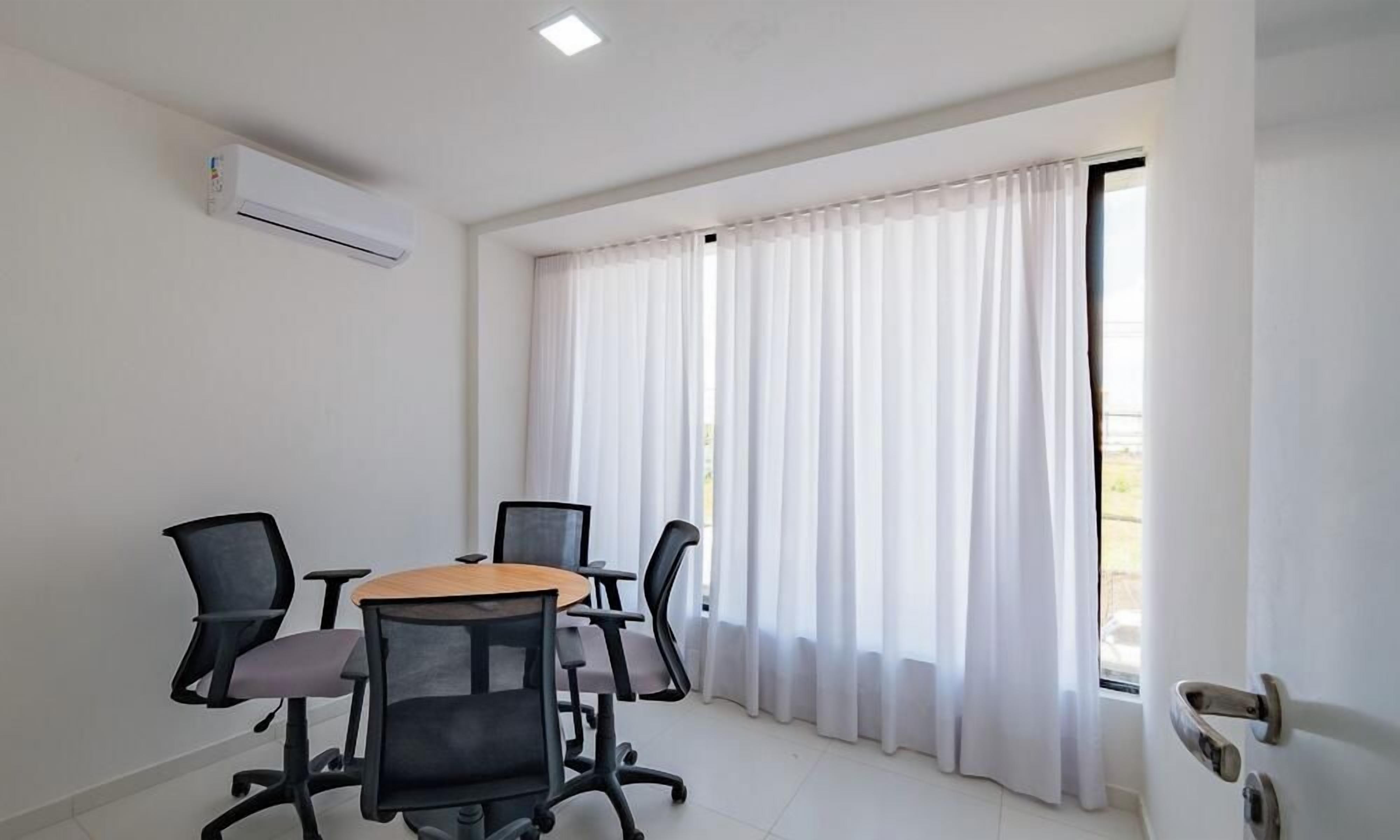 A conference room with a table, four chairs, a white air conditioning unit, and a window with white curtains.