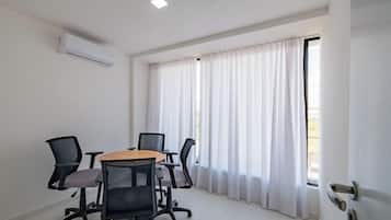 Uma sala de conferências com mesa, quatro cadeiras, um aparelho de ar condicionado branco e uma janela com cortinas brancas.