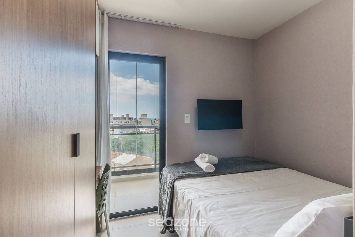 A bedroom with a bed, a TV mounted on the wall, and a view of the city through a large window.