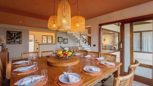 Un coin repas avec une table en bois dressée pour six personnes, entourée de chaises en osier, agrémenté d’un centre de table composé d’un panier de fruits et d’un luminaire suspendu.