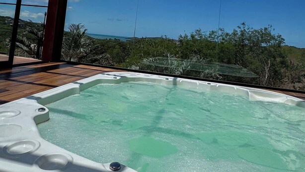 Un bain à remous dans un espace extérieur abrité, doté d’une terrasse en bois, entouré de verdure et surplombé d’un ciel bleu dégagé.
