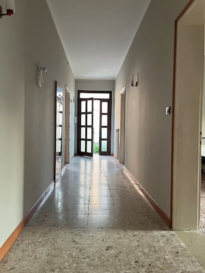 Un pasillo con piso de baldosas brillantes, una puerta al final y una ventana con vista a follaje verde.