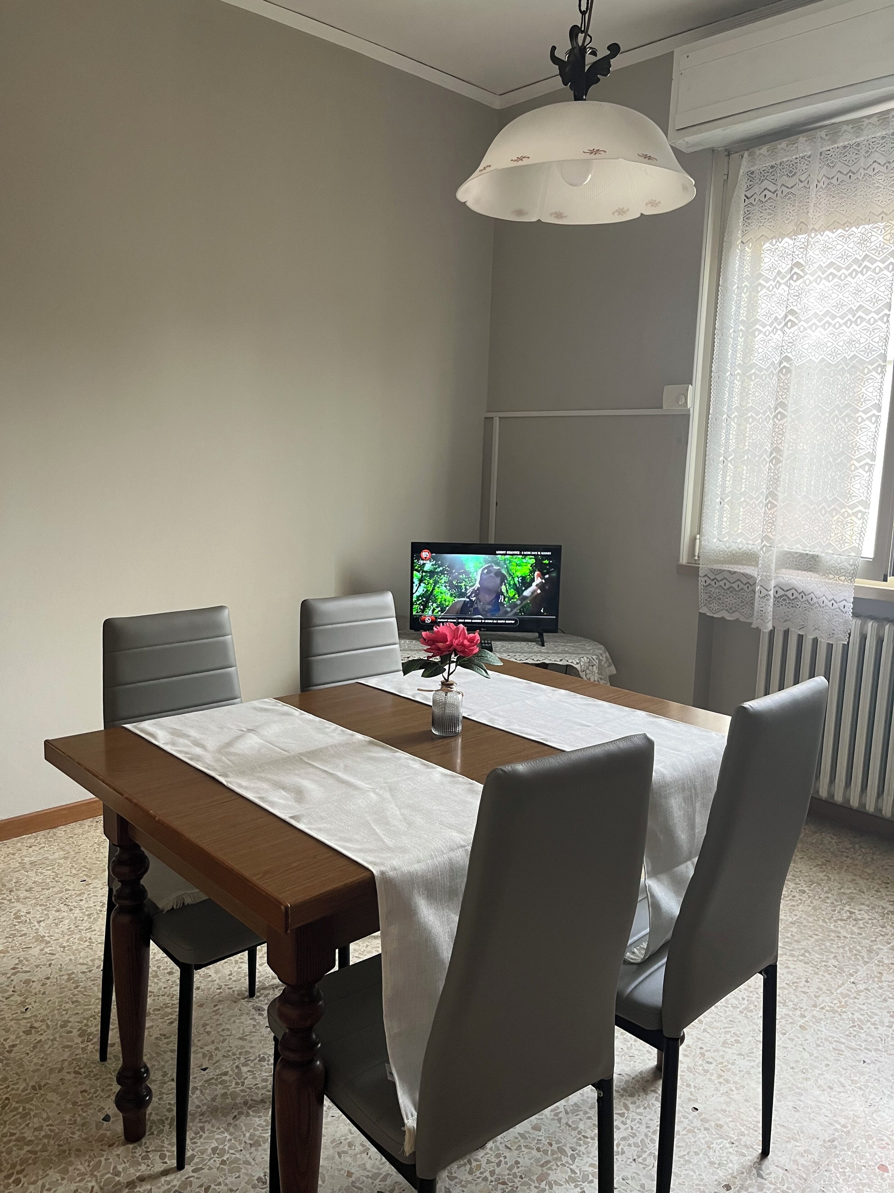 A dining area with a wooden table, white tablecloth, and grey chairs. A television is on in the background.