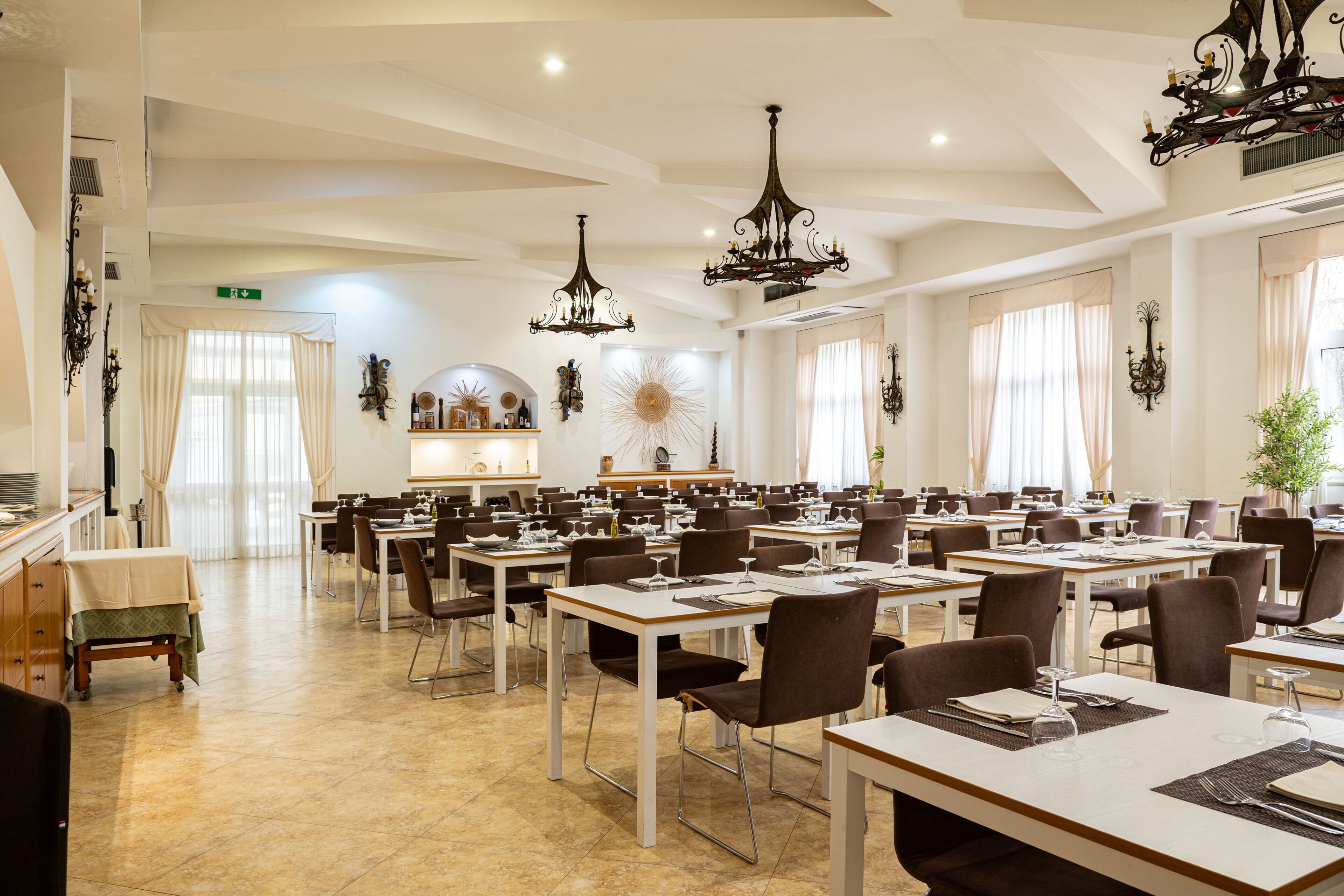 Un comedor amplio con varias mesas y sillas, candelabros elegantes y ventanas grandes con cortinas.