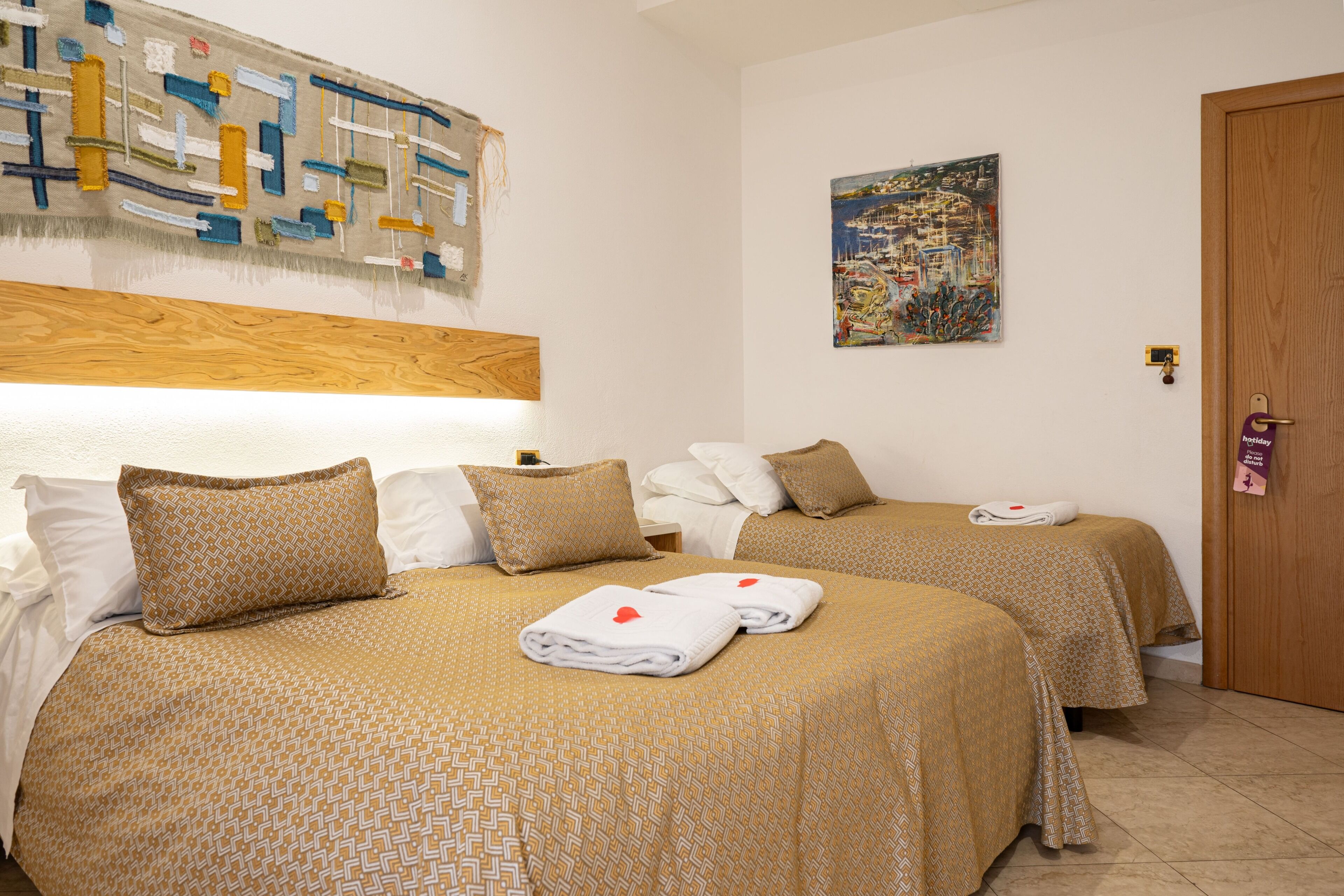 Habitación de hotel con dos camas, cada una con ropa de cama y toallas estampadas, cabeceros de madera, una obra de arte en la pared y una puerta con cerradura.