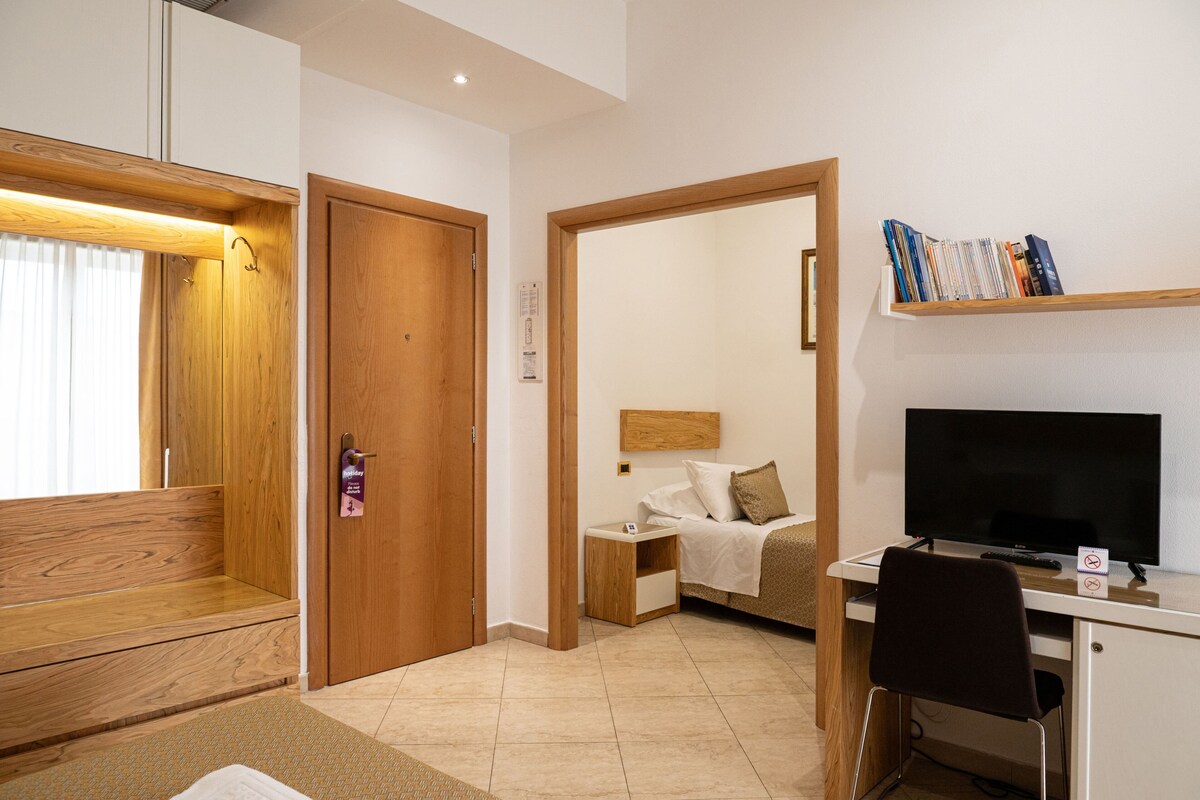A hotel room with a wooden bench, a bed with a headboard, a desk with a television, and a shelf with books.