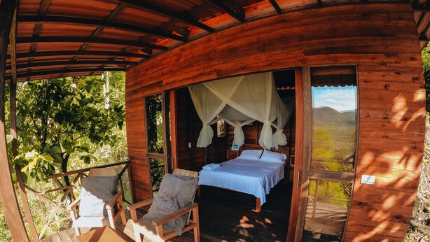 A wooden cabin with a bed, two chairs, and a table on a deck. - Nativa Ecohotel - Laguna de San Diego (Samaná)
